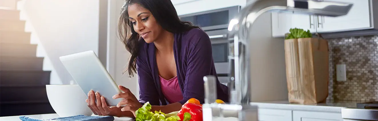 A woman standing in a kitchen, looking at a computer tablet.